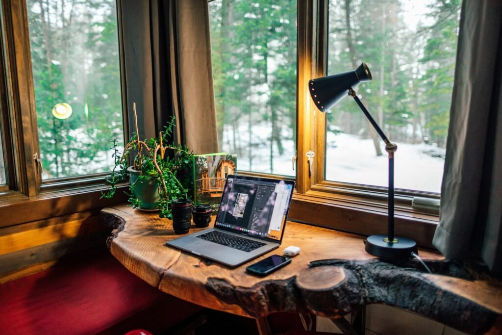 Laptop and phone on a live-edge wood desk by a cabin window overlooking snowy pines, workspace for handling hosting tasks